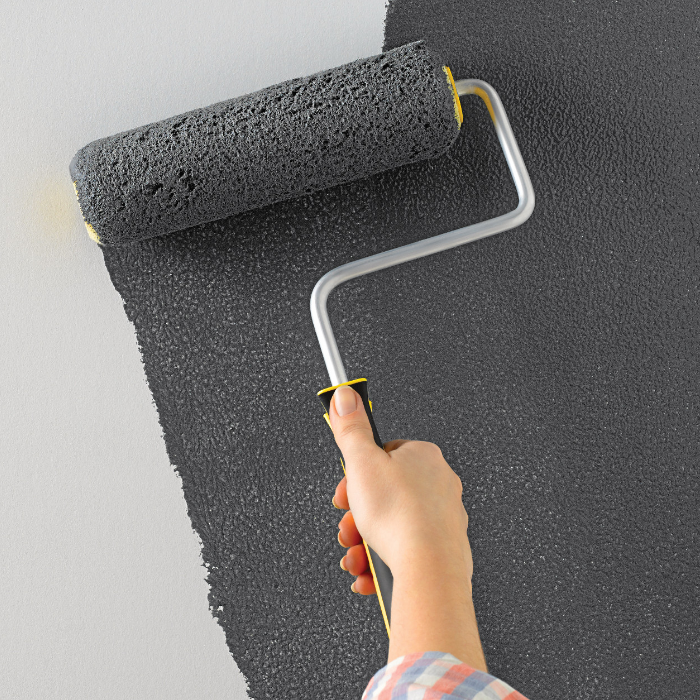 A woman applies dark grey paint to a white wall using a paint roller.